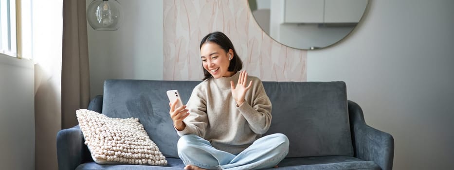 Portrait of asian woman sitting on sofa with smartphone, waving at mobile phone screen and waving at camera, video chat, talking to someone.