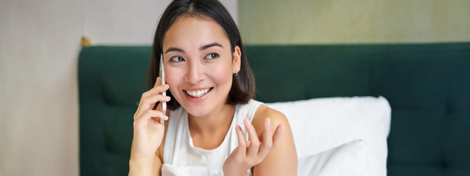 Close up portrait of cute asian girl in bed, talking on mobile phone with happy smiling face. Woman waking up and making a telephone call.