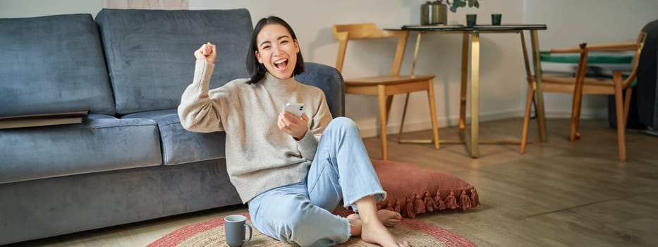 Enthusiastic asian woman sits on floor with smartphone, raise hand up and cheering, celebrating victory.