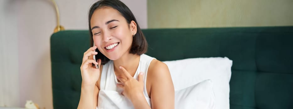 Close up portrait of cute asian girl in bed, talking on mobile phone with happy smiling face. Woman waking up and making a telephone call.