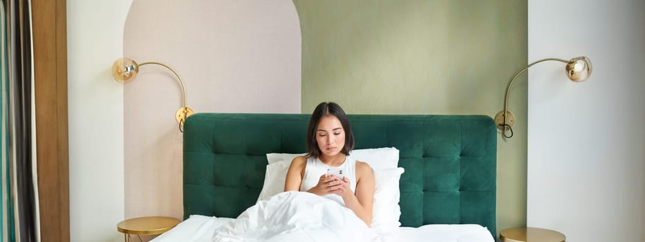 Portrait of woman lying in bed under duvet, looking at smartphone with serious face, reading message, watching news on mobile phone app.