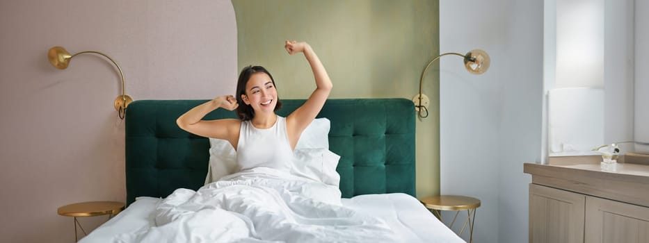 Beautiful asian woman lying in her bedroom on bed, stretching hands and looking outside, waking up from sleep.