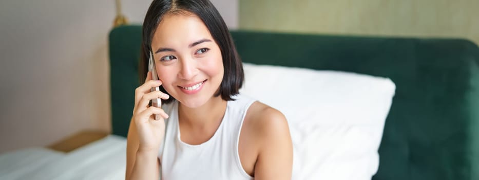 Smiling korean girl in bed, talks on mobile phone, making a phone call, lazy morning as asian woman orders delivery via smartphone.