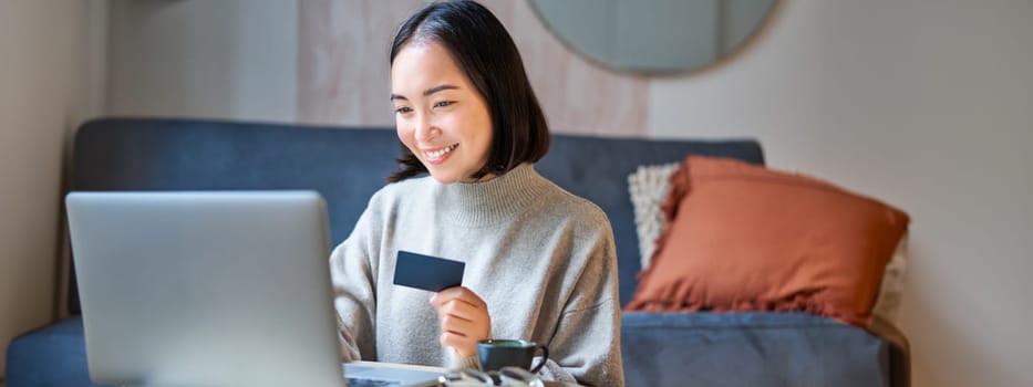 Portrait of korean woman shopping online, using her credit card and laptop to order delivery from website.