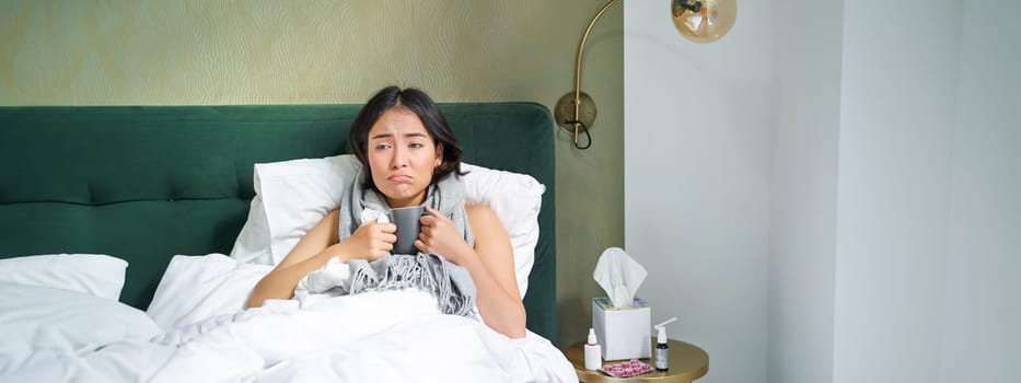 Sick sad asian girl lying in her bed with flu, catch a cold, taking pills and drinking hot tea, feeling upset.