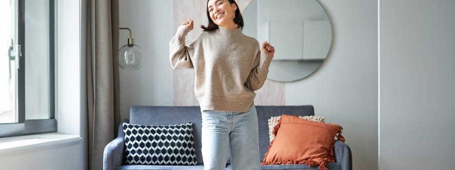 Enthusiastic asian woman dancing, feeling free and upbeat at her home, enjoying happy day, staying indoors, smiling and laughing.