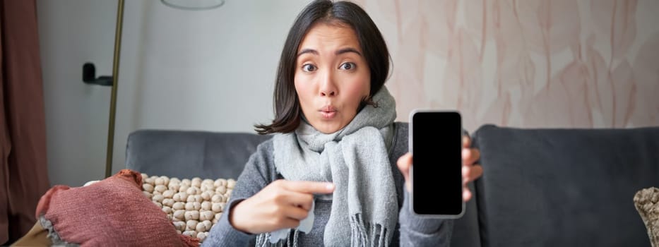 Excited young woman pointing finger at smartphone, showing online doctor, medical application or GP contact on mobile phone, staying at home sick, catching cold.