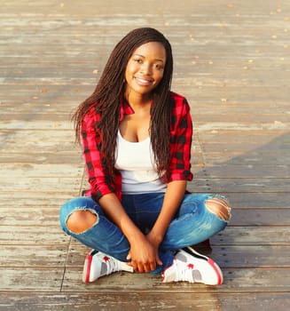 Fashion pretty young african woman sitting in the city park, wearing a red checkered shirt and cap 