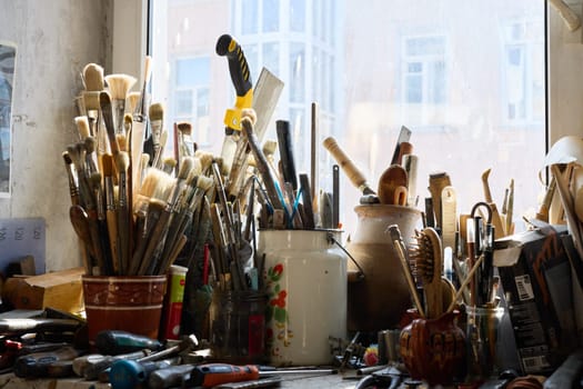A cluttered workspace features an array of brushes, tools, and containers, illuminated by natural light from a nearby window, reflecting an artist's vibrant creativity.