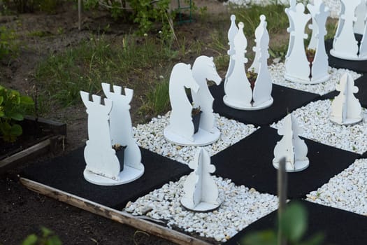A large and beautifully designed white chess set, featuring unique pieces, is set amidst lush, vibrant greenery