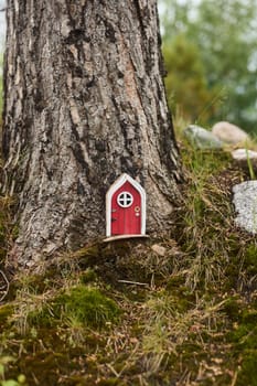A charming and whimsical miniature red door is nestled at the very base of a tree, surrounded by lush greenery