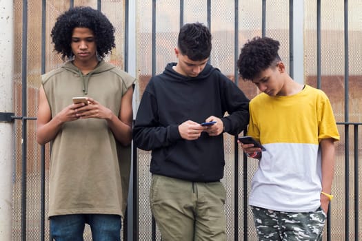 three serious multiracial teen friends browsing Internet on mobile phones standing against metal fence and looking at screen, concept of youth and technology of communication