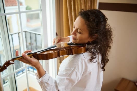 A woman smiles gently as she plays the violin by a sunlit window. The warm ambiance and her focused expression convey a sense of peace and creativity, capturing a moment of musical inspiration.