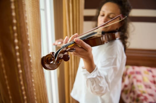A woman gracefully plays the violin near a sunlit window, creating a serene and intimate atmosphere. The warm colors and soft curtains add to the cozy ambiance, reflecting a moment of musical peace.