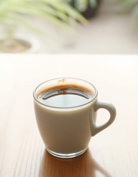A clear glass mug filled with rich, dark coffee is placed on a light wooden table. Gentle sunlight filters through a nearby window, creating a warm atmosphere.