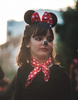 The image shows a child dressed in a costume that includes what looks like Mouse ears, commonly associated with the iconic Disney character Minnie Mouse. The child appears to have a red bow with white polka dots, which is Minnie Mouse's signature style. Additionally, there is face paint resembling a small black nose and whiskers, contributing to the mouse-themed appearance. The child is also wearing a red scarf with polka dots, matching the bow on top of the head. The background is blurred, putting the focus on the child, who seems to be looking off into the distance.