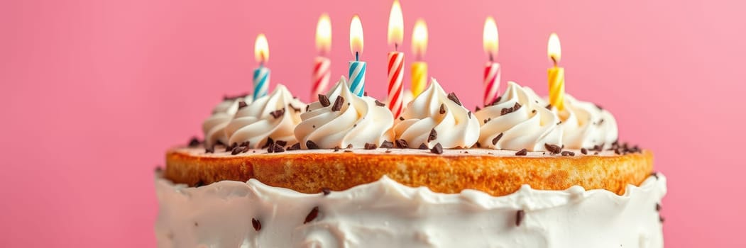 A birthday cake with creamy frosting topped with colorful sprinkles and lit candles stands against a vibrant pink backdrop. This delightful dessert is ready for a celebration.