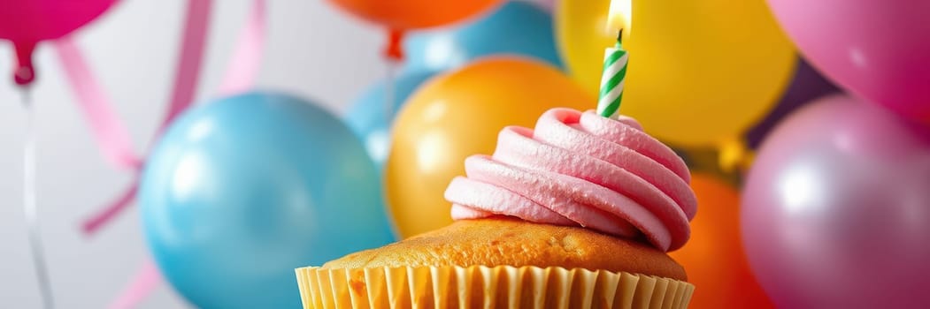 A delicious cupcake topped with pink frosting and a lit candle sits in front of vibrant balloons of various colors, celebrating a joyful birthday occasion.