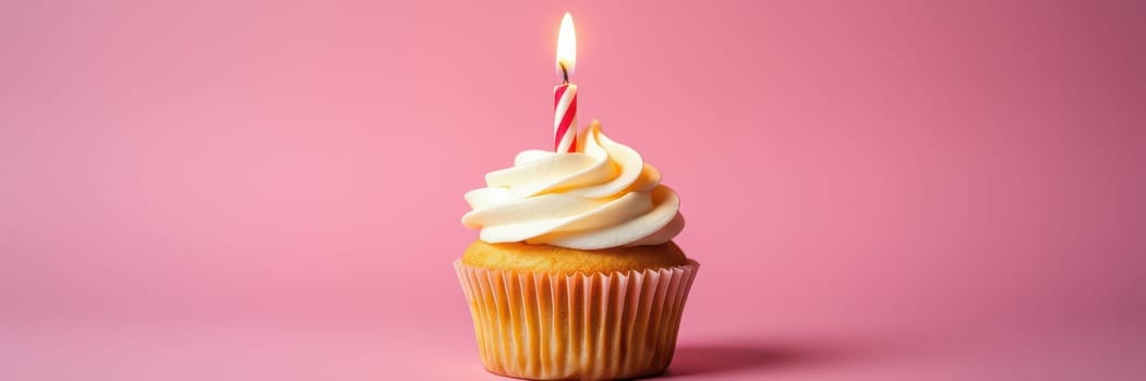 A vanilla cupcake sits on a table adorned with swirls of creamy frosting and a lit candle. The vibrant pink background enhances the festive atmosphere perfect for a birthday.