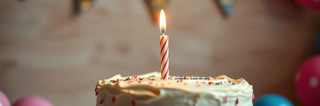 A festive cake with a single lit candle on top, adorned with colorful sprinkles, is the centerpiece of a birthday celebration decorated with balloons in vibrant colors.