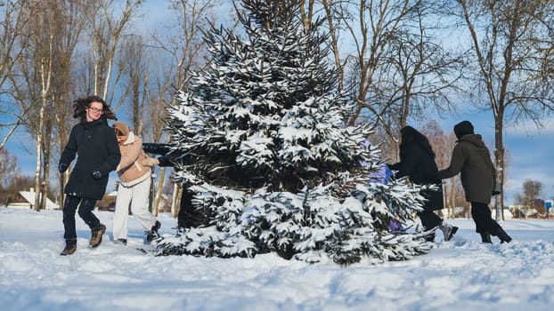 Cheerful schoolgirls are enjoying winter holidays playing hide and seek around a snow covered fir tree in a city park