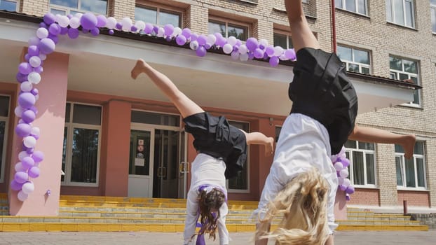 Two schoolgirls performing cartwheels in front of their school on the final day of school, marking the occasion with purple and white balloons
