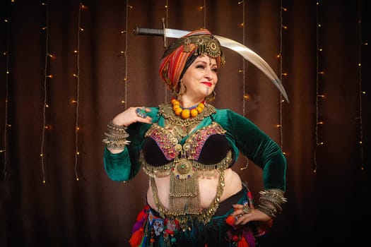 A woman in a colorful headscarf and jewelry poses With a saber in front of wall of colorful tassels