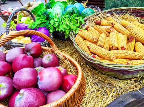 Colorful baskets filled with apples, corn, and leafy lettuce create a vibrant display at a bustling market in fall.