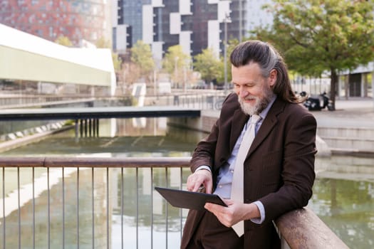 positive middle age male with beard in formal outfit browsing tablet while standing at city, concept of business and mature man lifestyle, copyspace for text