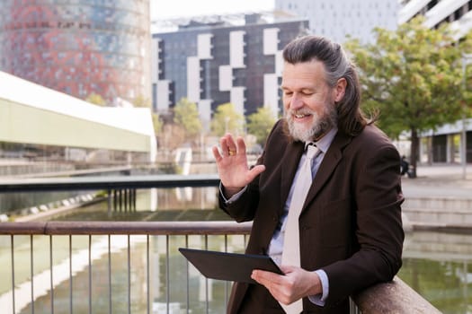 happy middle age male with beard in elegant suit waving hand while greeting partner during video meeting via digital tablet on terrace, concept of business and mature man lifestyle, copyspace for text