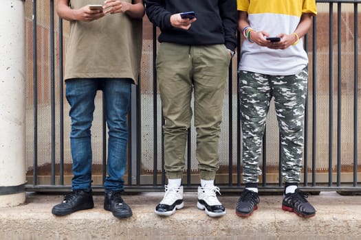 crop of three unrecognizable teen friends browsing Internet on mobile phones standing against metal fence and looking at screen, concept of youth and technology of communication