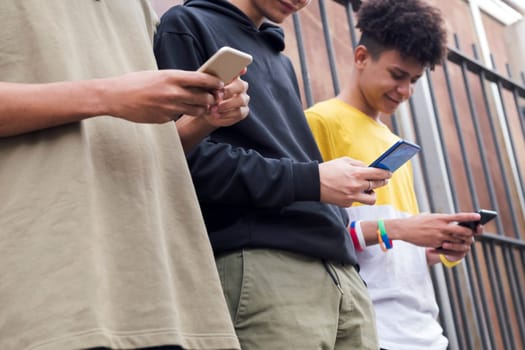 crop of three unrecognizable teen friends browsing Internet on mobile phones standing against metal fence and looking at screen, concept of youth and technology of communication