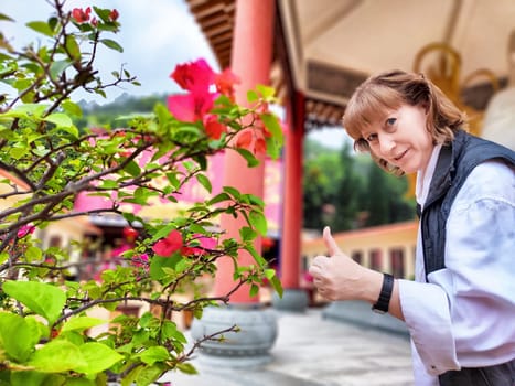 Blonde woman gives a thumbs up in a vibrant temple garden filled with colorful flowers during her travel excursion.