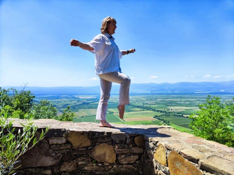 A middle-aged woman balances on a stone wall, enjoying a scenic view of fields and mountains on a bright, sunny day.