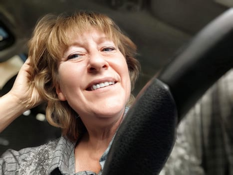 A middle aged woman with a cheerful expression is sitting in her car, gently touching her hair and enjoying the sunny afternoon.