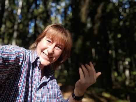 A middle-aged woman is taking a selfie in the forest, smiling happily while exploring nature and sharing her travel experiences.