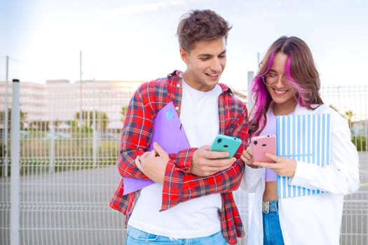 A young couple enjoys using their smartphones while standing outdoors. They smile at each other, holding colorful notebooks. The background shows a sunny day and a fence, creating a relaxed vibe.