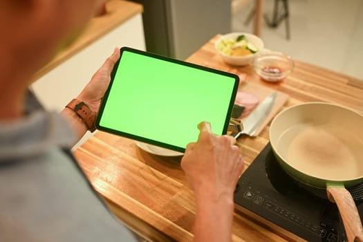 Close up of senior man using digital tablet in a cozy home kitchen to follow a recipe or cooking instructions.