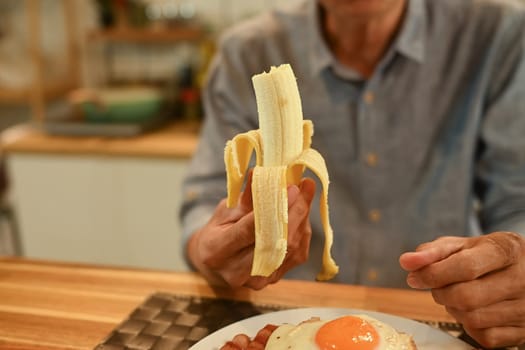Senior man holding peeled banana while enjoying breakfast at kitchen table..