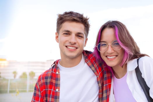 happy young couple smiles together in an outdoor setting. The woman has pink highlights in her hair and wears glasses, while the man sports a plaid shirt. A fence and greenery create a relaxed vibe