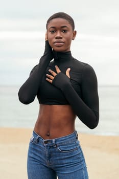 young gentle african american woman in casual clothes touching cheek while looking at camera on ocean coast