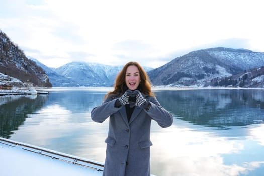 A happy woman in mittens and a coat forms a heart with her hands, enjoying the serene snowy fjord and mountain views in Gaupne, Norway. 
