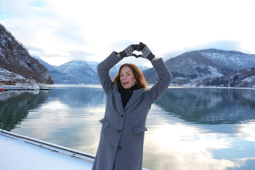 Happy woman in a coat and mittens forms a heart with her hands by a serene snowy fjord and mountains in Gaupne, Norway