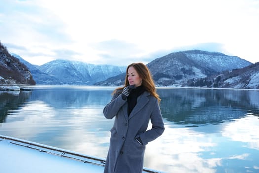 A serene woman in a coat and mittens enjoys the peaceful snowy fjord and mountain views in Gaupne, Norway, during winter. 