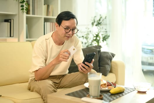 Smiling senior man using a credit card for online payment with mobile phone at home.