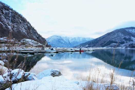 A tranquil winter marina in Gaupne, Norway, with boats docked on calm waters, surrounded by snowy mountains and stunning reflections