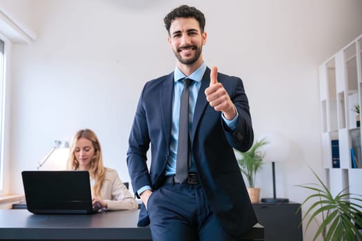 A confident businessman gives a thumbs up while standing in the office. His colleague works on a laptop in the background. The scene reflects a positive and productive work environment.