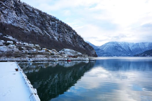A scenic snowy village in Gaupne, Norway, overlooking a calm fjord surrounded by majestic mountains and serene winter reflections