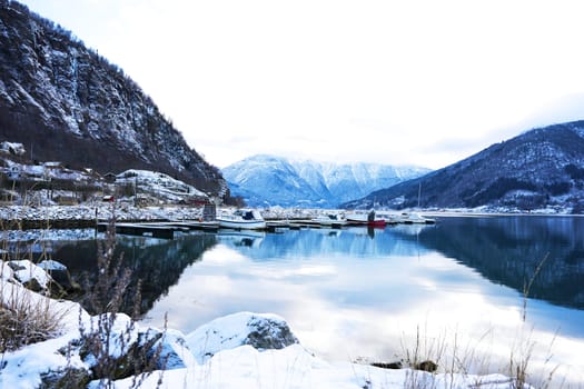 A scenic winter marina in Gaupne, Norway, with snow-covered mountains, calm waters, and boats docked along the fjord's edge. 