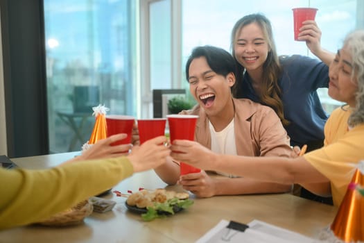 Group of colleagues laughing and clinking cups to celebrate at office party.
